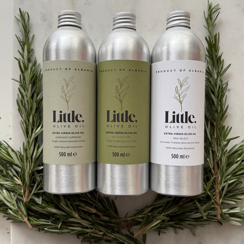 Three silver aluminum bottles of Little Olive Oil arranged side by side on a light marble surface, framed by green rosemary sprigs.