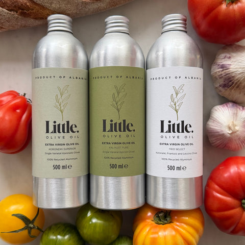 Three silver aluminum bottles of Little Olive Oil arranged in a styled flat-lay with tomatoes, garlic, and peppers around them. 