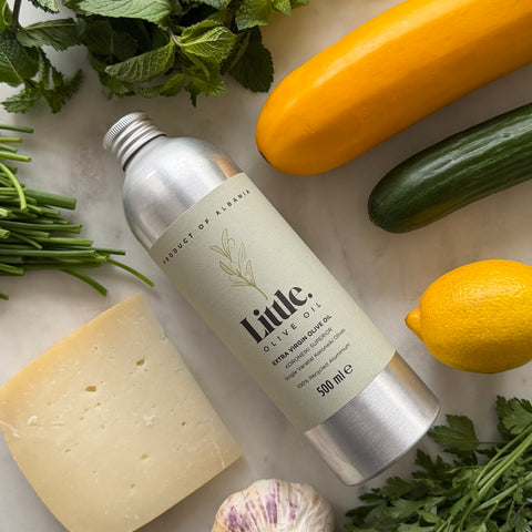 A bottle of Little Olive Oil Koroneiki Superior Extra Virgin Olive Oil surrounded by fresh ingredients including parsley, garlic, cheese, yellow squash, cucumber, and lemon on a light marble surface.