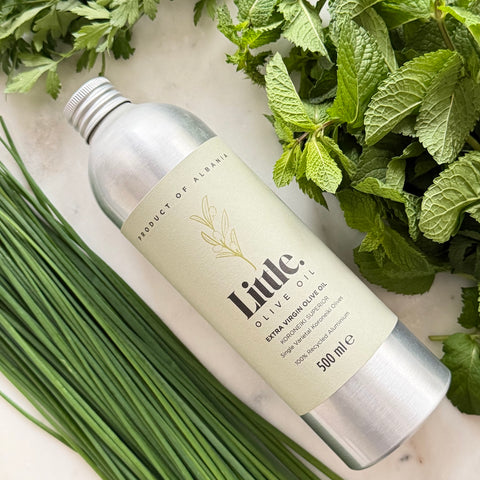 A 500 ml bottle of Little Olive Oil Koroneiki Superior extra virgin olive oil, styled in a flat-lay with fresh green herbs (mint, parsley, chives) on a light marble background.