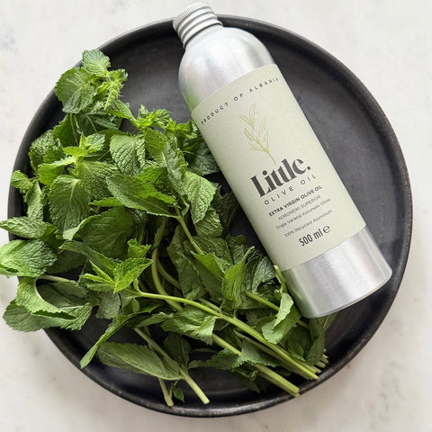 A 500 ml bottle of Little Olive Oil Koroneiki Superior sits on a black plate next to a generous pile of fresh green mint leaves, set against a light marble background.