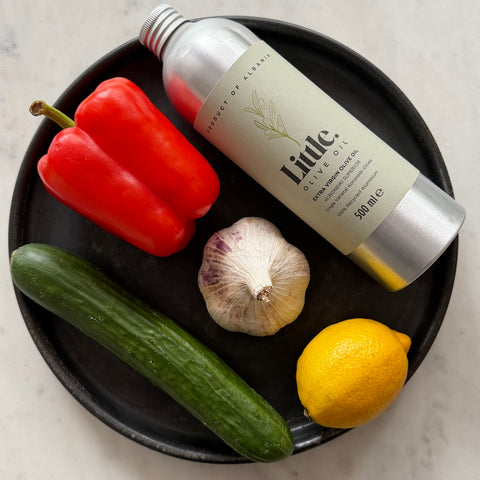 A 500ml bottle of Little Olive Oil Koroneiki Superior with ingredients arranged on a black plate with a red bell pepper, cucumber, garlic, and lemon on a light marble surface.