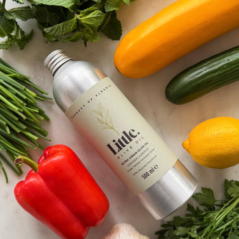 A 500 ml bottle of Little Olive Oil positioned diagonally on a light marble surface, surrounded by fresh ingredients including a red bell pepper, green cucumber, yellow squash, lemon, mint, chives, and parsley.