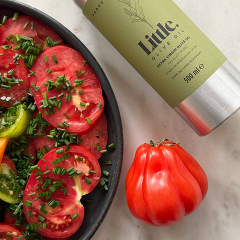 A bottle of Little Olive Oil extra virgin olive oil lies on a light marble surface next to a black bowl filled with sliced tomatoes, chive, and other vegetables, with a whole red tomato in front.