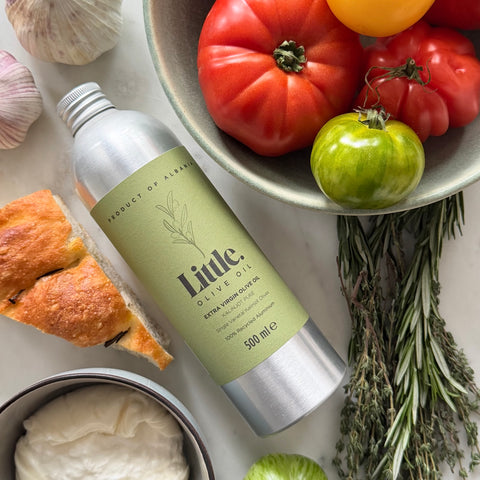 A bottle of Little Olive Oil Extra Virgin Olive Oil lies on a light marble surface surrounded by food props including tomatoes, garlic, bread, cheese, and rosemary.