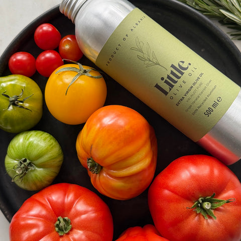 A 500 ml bottle of Little Olive Oil with a light olive-green label and silver cap, styled on a black plate with fresh tomatoes.