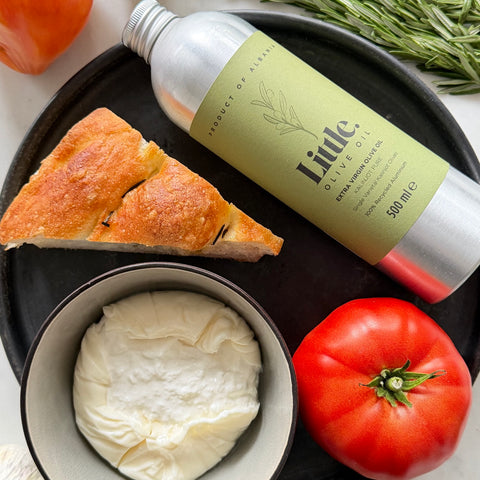 A bottle of Little Olive Oil extra virgin olive oil displayed on a black plate with a slice of bread, a bowl of creamy cheese, and a red tomato.