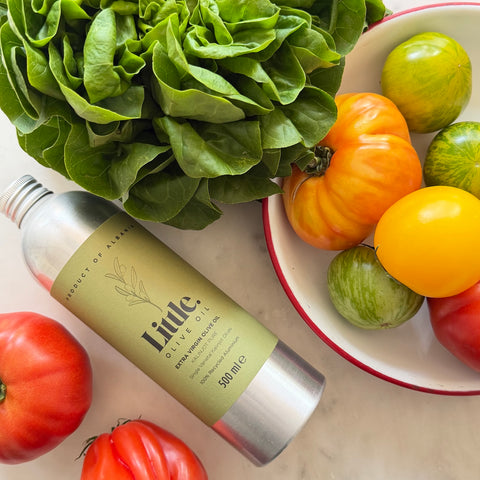 A bottle of Little Olive Oil placed on a light marble surface with fresh tomatoes and a bowl of mixed tomatoes around it. The bottle label reads 'Product of Albania' and 'Little Olive Oil'.