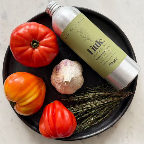 A silver metal bottle of Little Olive Oil with a light olive-green label, arranged on a black plate with tomatoes, garlic, and herbs on a light marble surface.