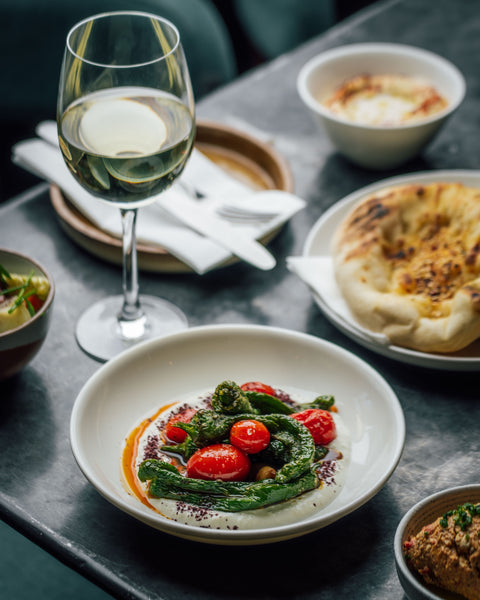 Dinner table with a dish of greens and tomatoes, a glass of white wine, and bread.