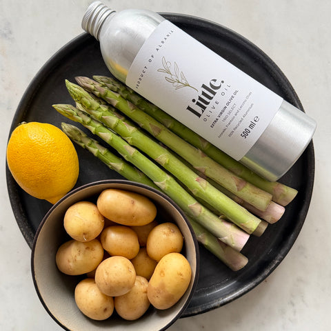 A bottle of Little Olive Oil styled on a black plate with fresh asparagus, a lemon, and a bowl of potatoes on a light marble surface.