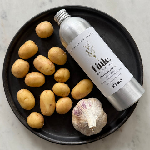 A 500 ml bottle of Little Olive Oil styled on a black plate with small potatoes, a garlic bulb, and a white marble surface.