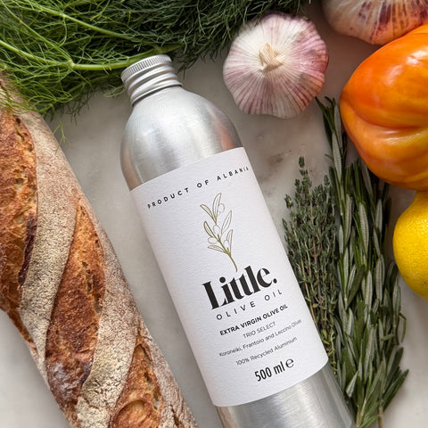 A 500 ml bottle of Little Olive Oil Extra Virgin Olive Oil from Product of Albania, styled in a flat-lay with fresh ingredients including a loaf of bread, garlic, a tomato, a lemon, and herbs on a light marble background.