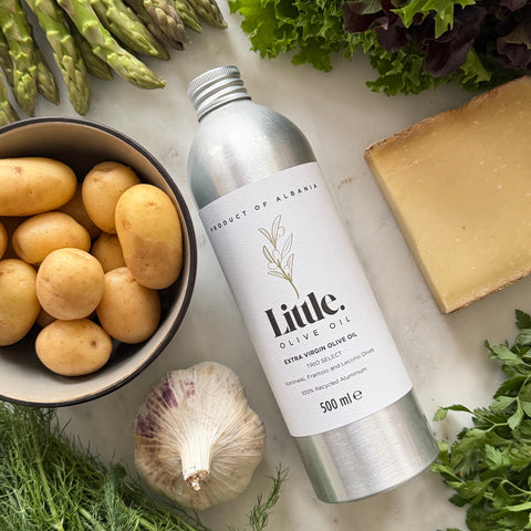 A 500 ml silver aluminum bottle of Little Olive Oil extra virgin olive oil, surrounded by fresh ingredients like asparagus, potatoes, garlic, cheese, and herbs on a light marble surface.